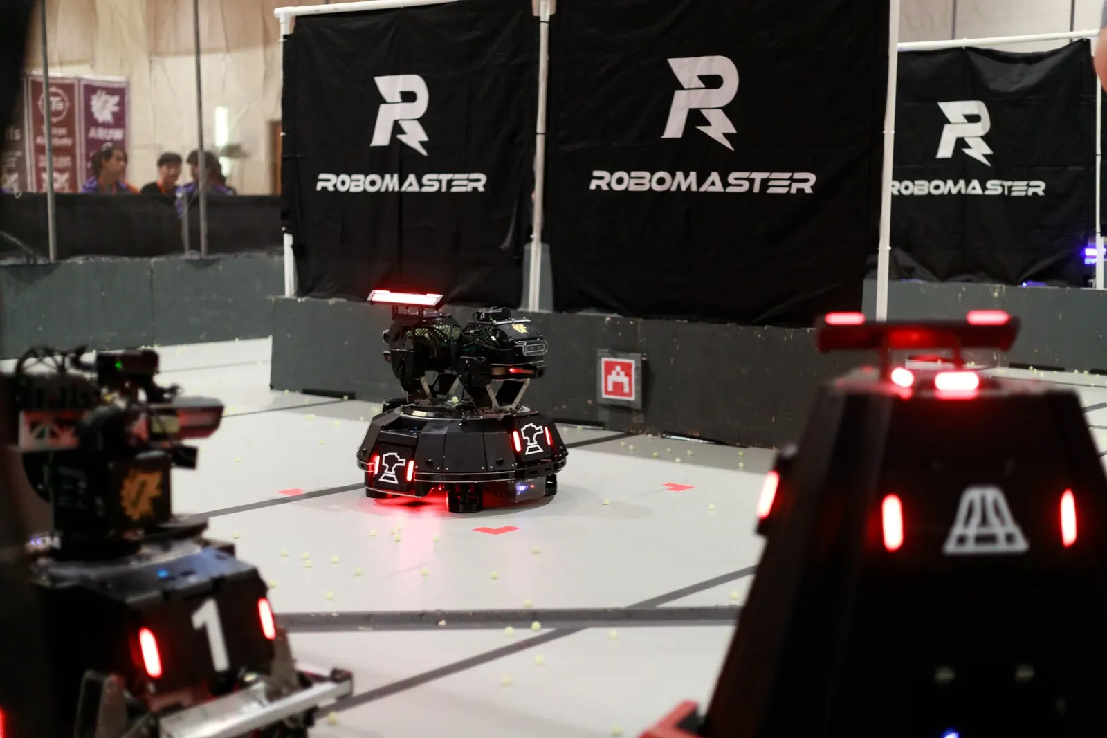 Robots with red LED armour facing off on the arena floor, spectators behind the barrier.